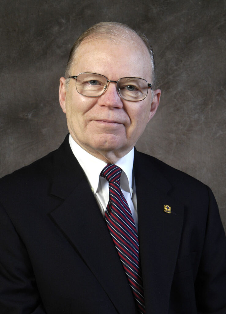 An older man wearing glasses, a dark suit, a white shirt, and a striped tie, poses in front of a brown textured background.