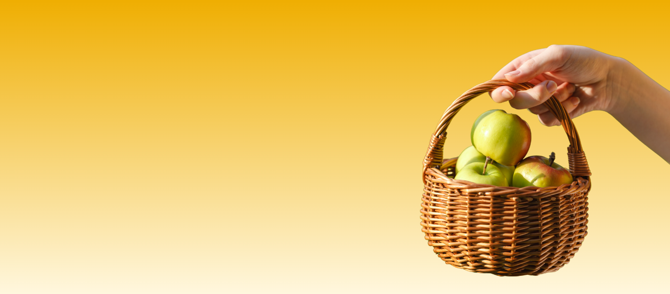 A hand holds a wicker basket containing several green apples against a yellow gradient background.