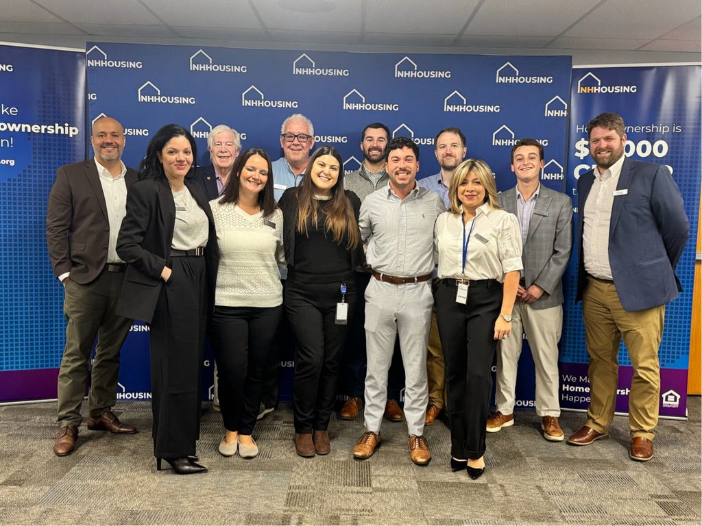 Twelve people stand posing and smiling in front of a blue NH Housing banner in a professional setting.