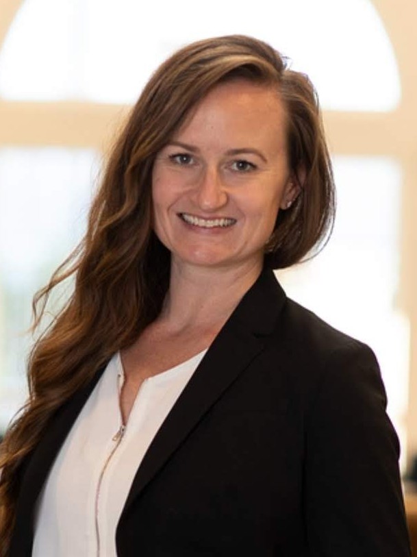 Woman with long hair stands indoors, smiling, wearing a black blazer, white shirt, and white pants. Bright window and chairs are visible in the background.