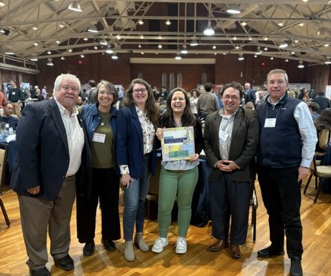 NH Housing staff and local partners stand in a large room with a wood floor, one woman in the center holding the award for the HOP Grant.